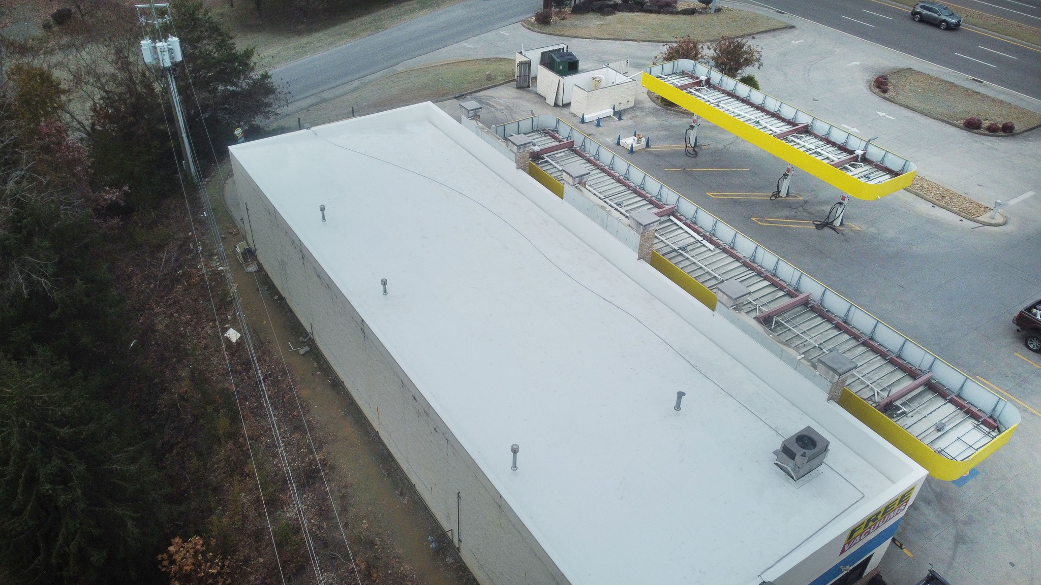 Aerial view of a flat-roofed commercial building next to a self-serve car wash area with yellow trim, bordered by roads and some trees.