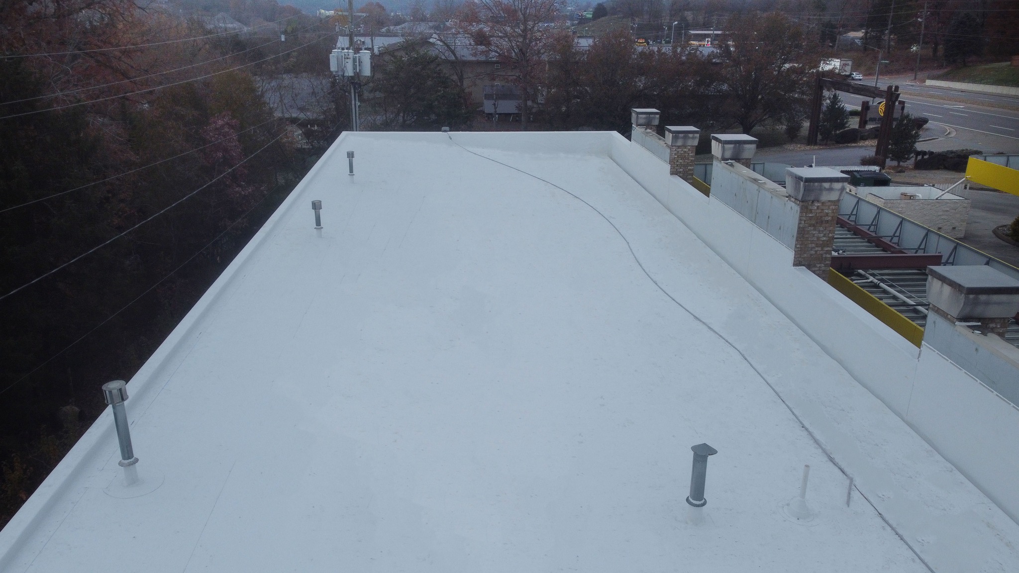 Flat white commercial building roof with multiple vent pipes and a visible cable or seam running across the surface, surrounded by trees and nearby roads.