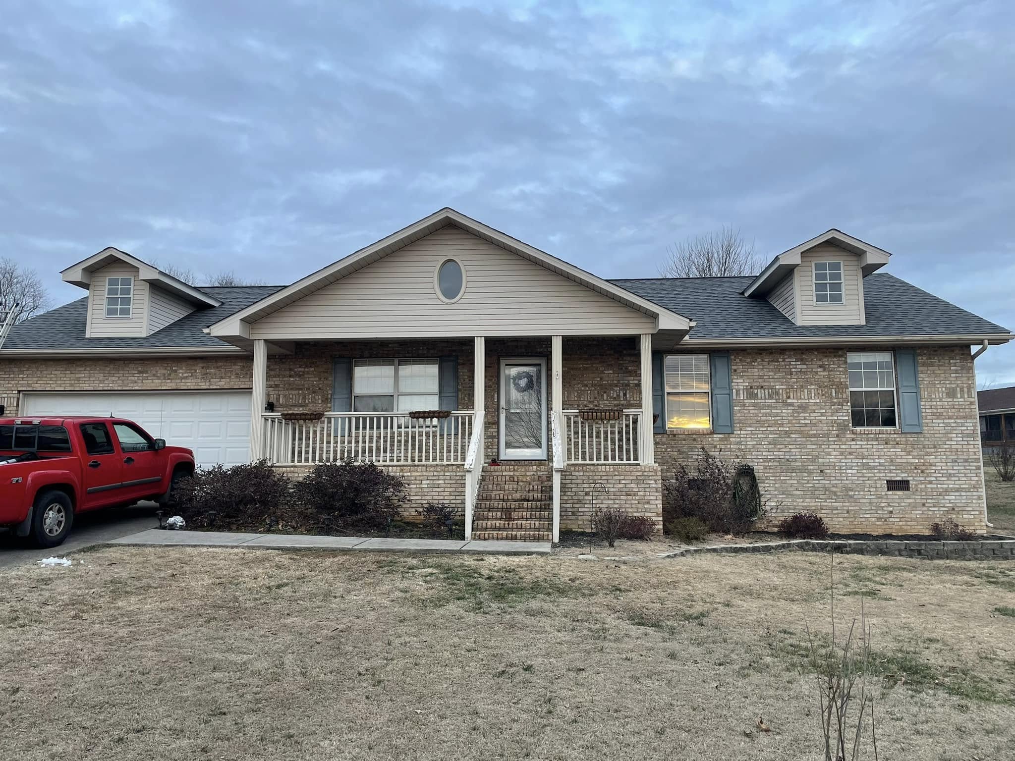 A single-story brick house with a covered front porch, two dormer windows, and a red pickup truck parked in the driveway on the left side.