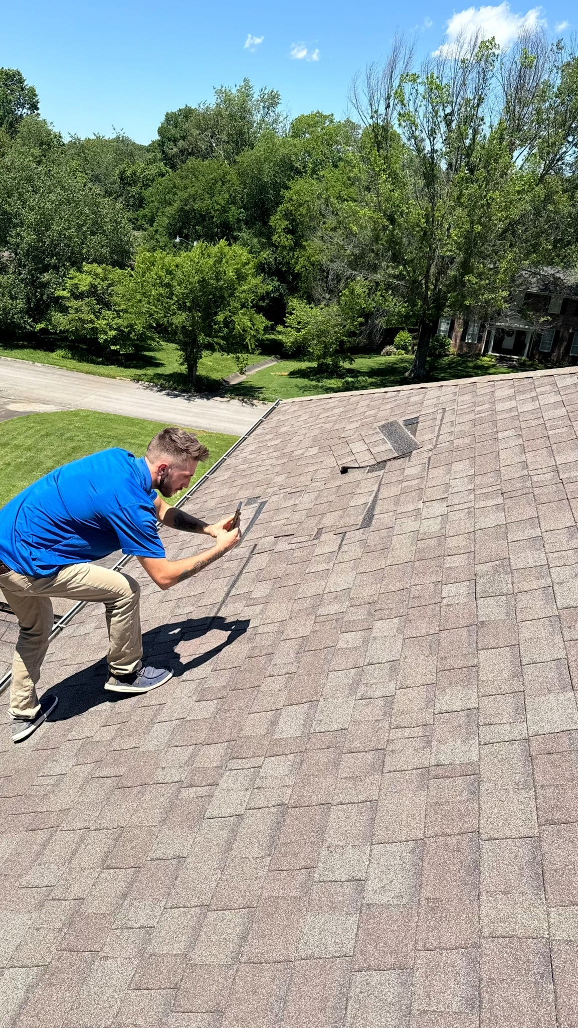 A person in a blue shirt takes a photo of damaged shingles on a house roof under a sunny sky, with trees and a road in the background.