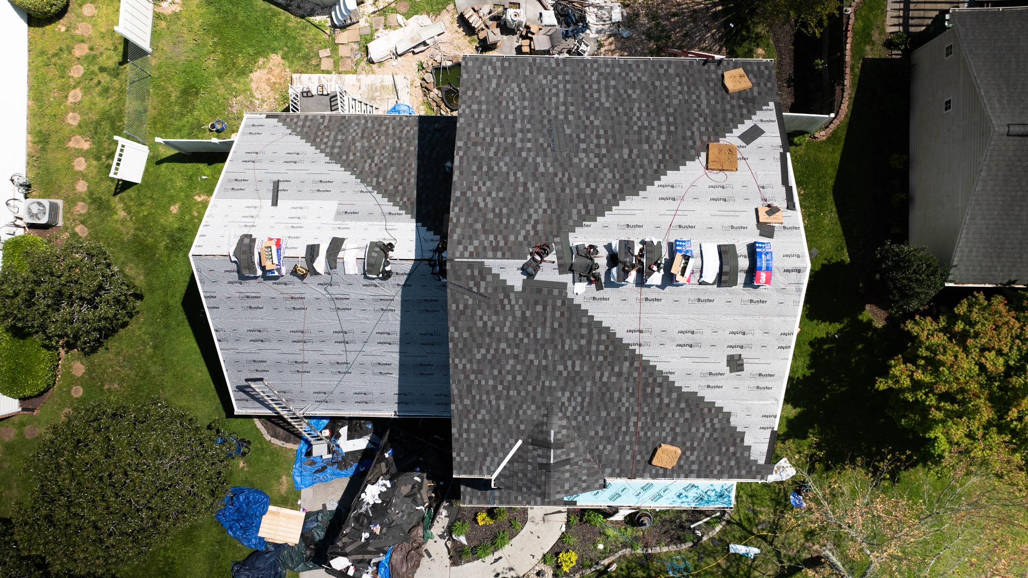 Aerial view of a house undergoing roof replacement, with workers, roofing materials, and ladders visible on the partially shingled roof.