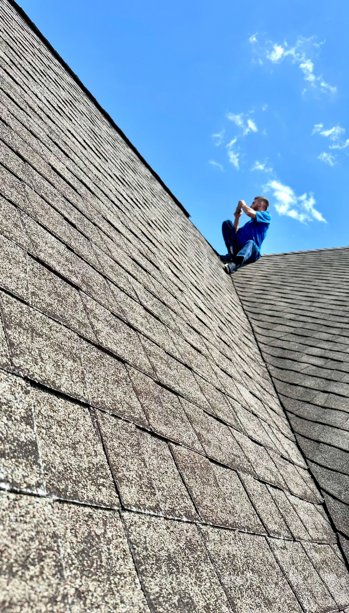 A person in a blue shirt sits near the peak of a shingled roof, holding a tool and working, with a clear blue sky in the background.