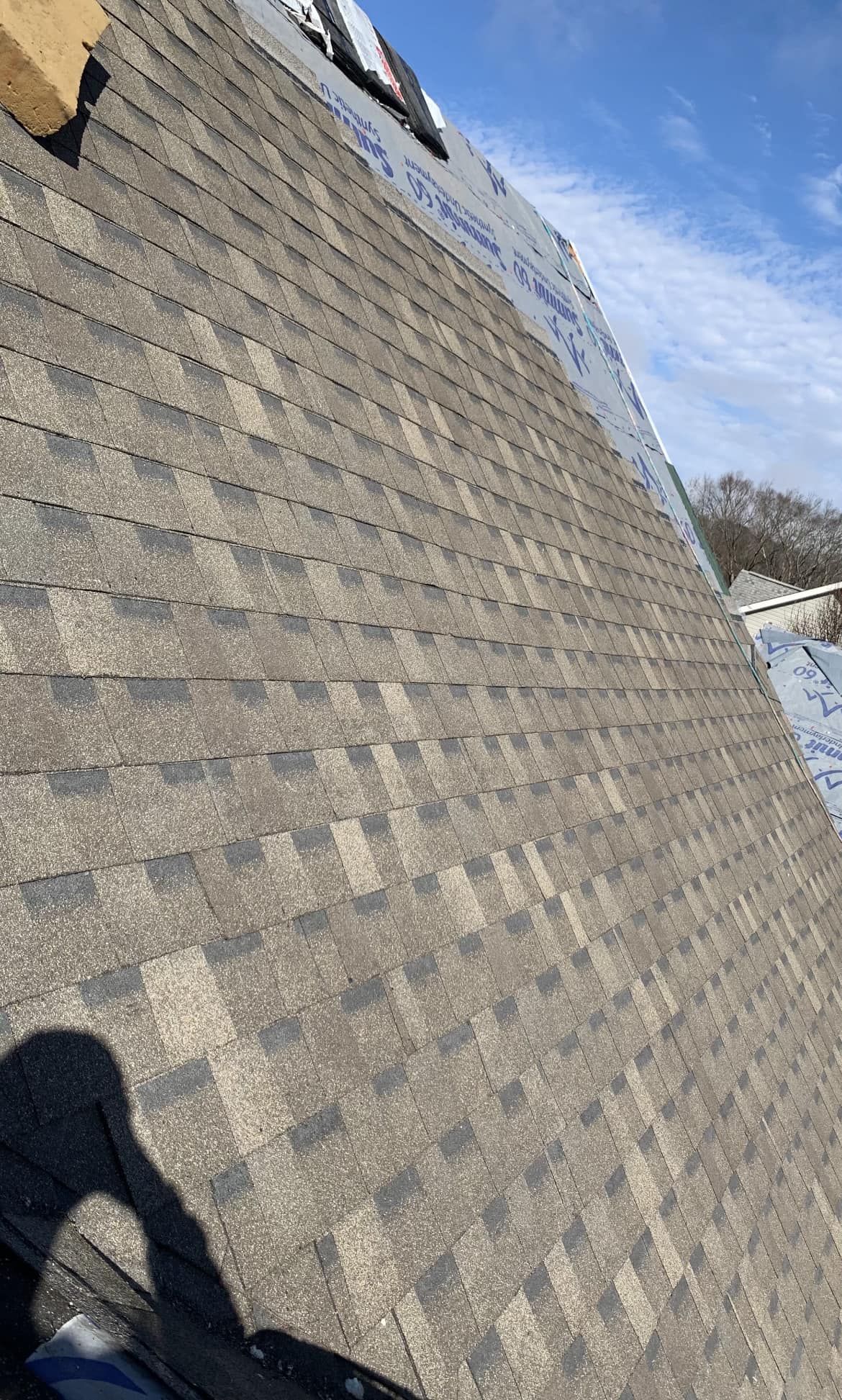 Sloped roof with new asphalt shingles partially installed, roofing materials visible at the ridge, and a clear sky in the background.